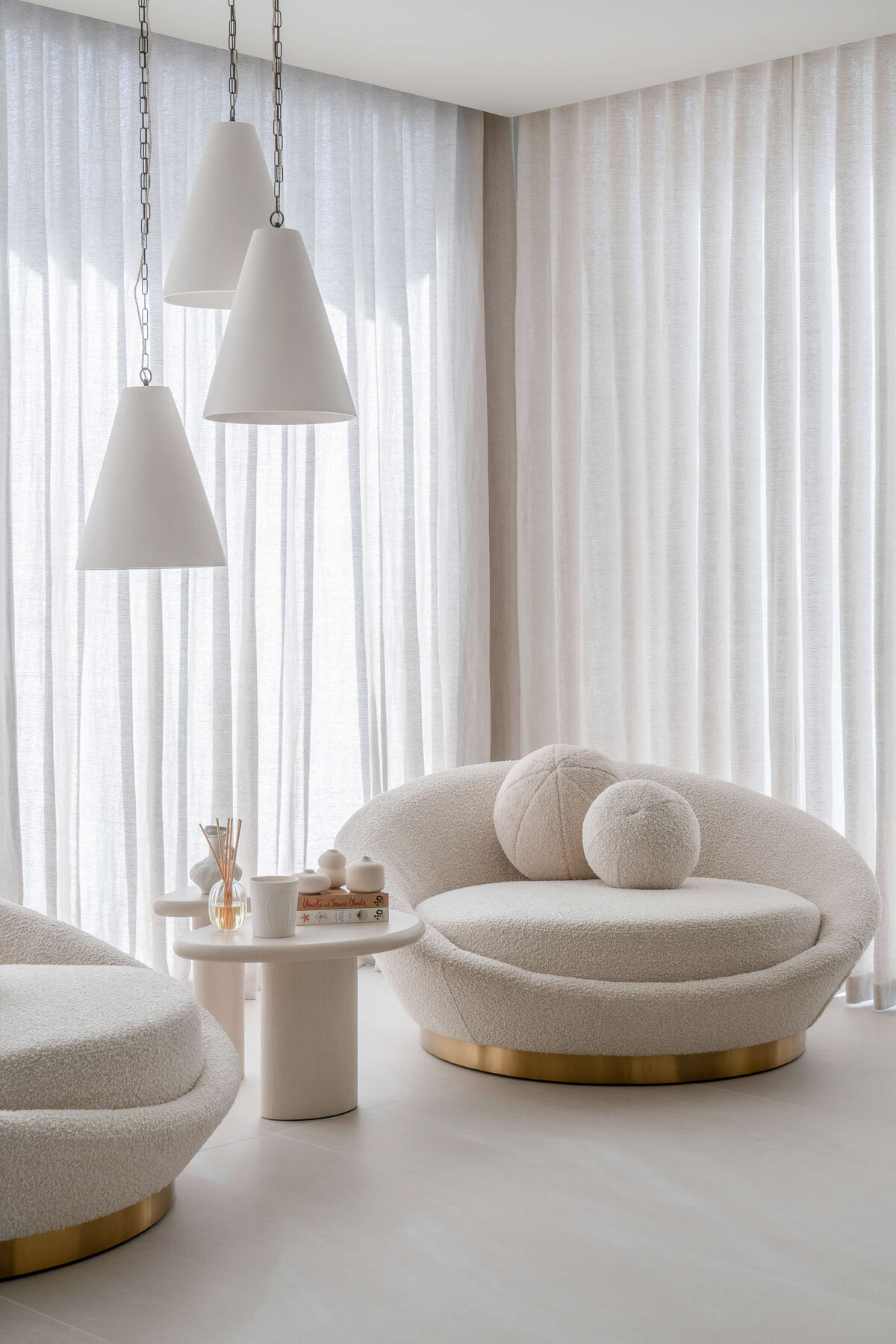 Two large round chairs with spherical pillows and shiny gold bases. Soft white curtains in the background and three white pendant lights overhead.