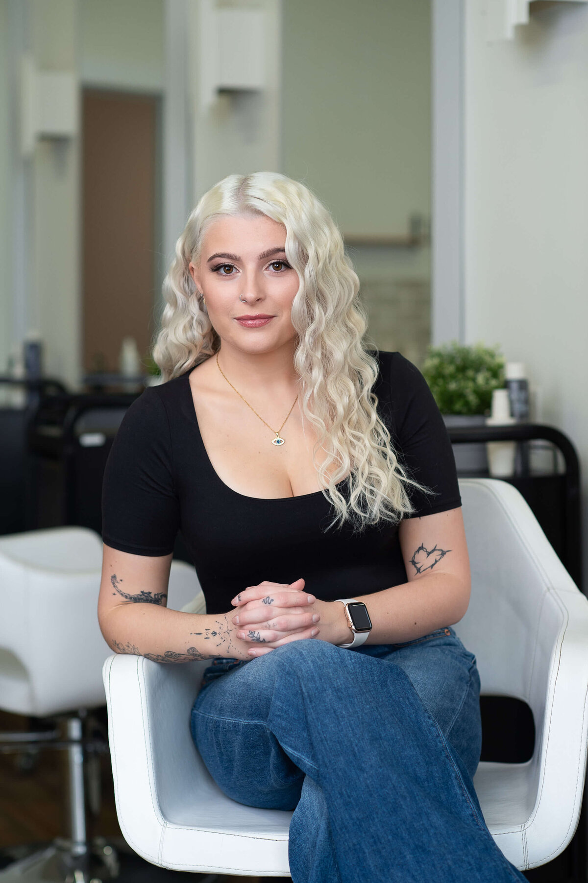 a profile photo of a young blonde hair stylist wearing a black top and jeans sitting in a white chair.  Captured on-location at Richardson Hair Design by Ottawa branding photographer JEMMAN Photography