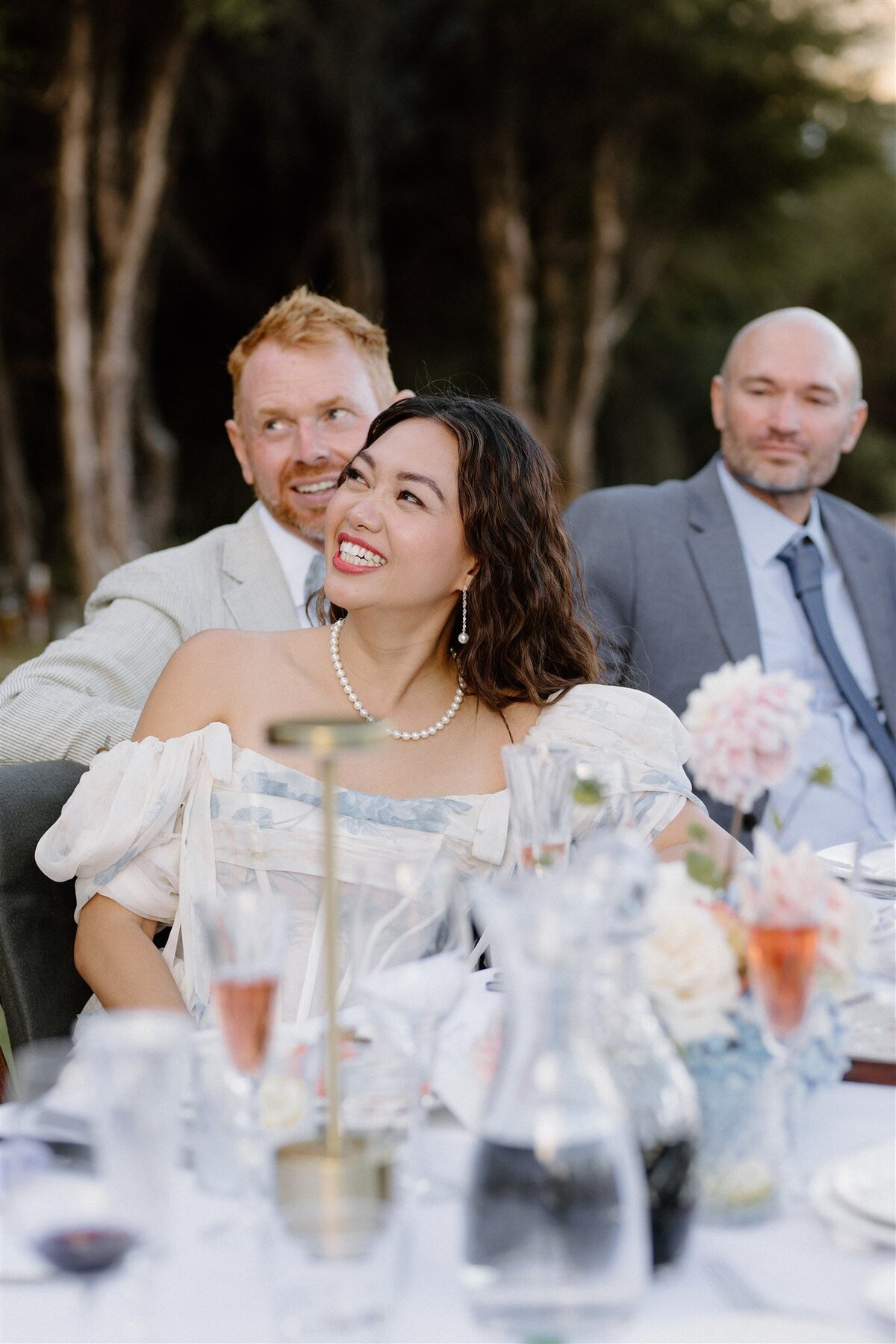 A smiling bride wearing a pearl necklace and drop earrings, seated beside her groom at a beautifully set outdoor table, listening to speeches.