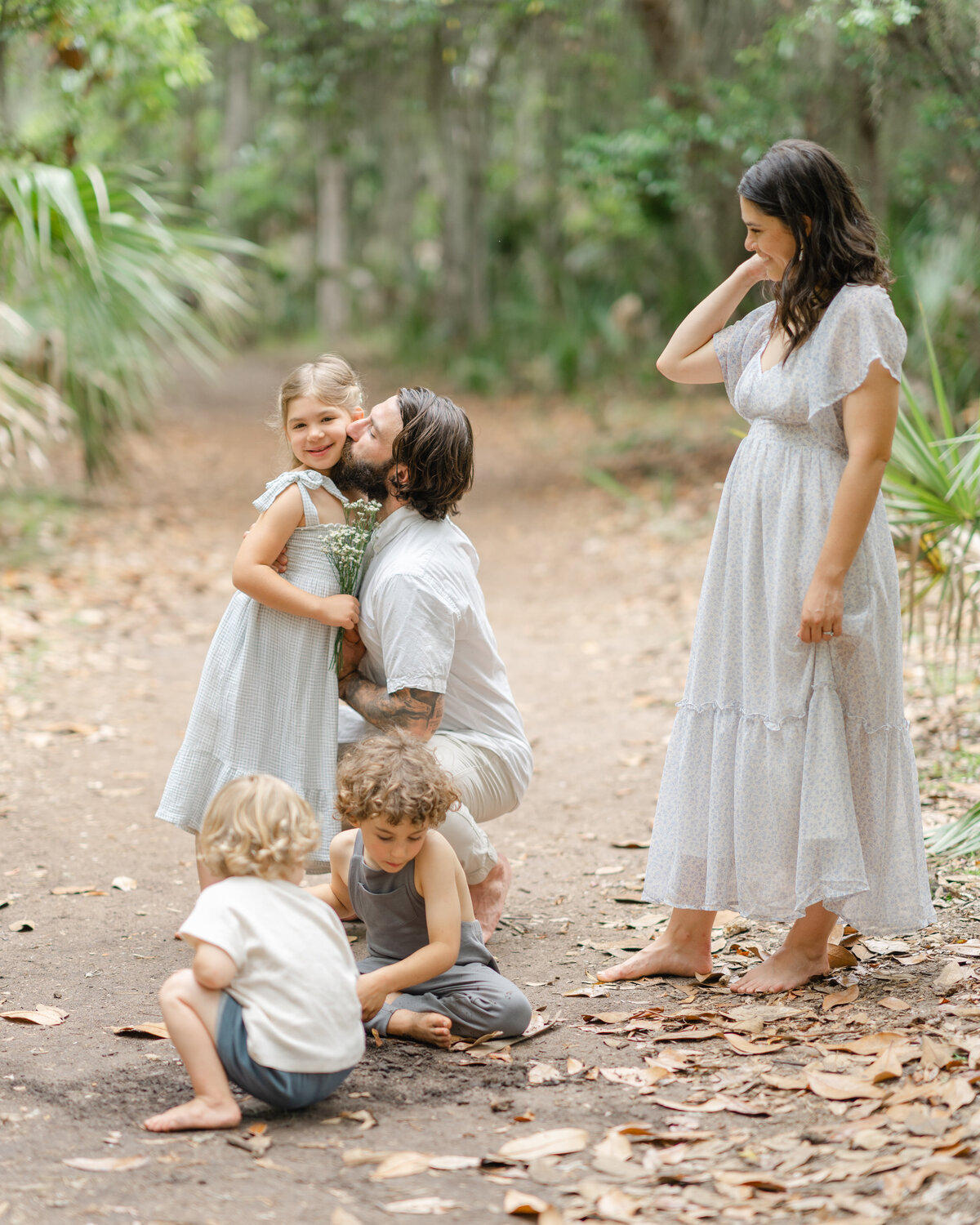 Father kisses daughters cheek as brothers play in the sand on a path in Wormsloe plantation by Savannah family photographer Courtney Cronin