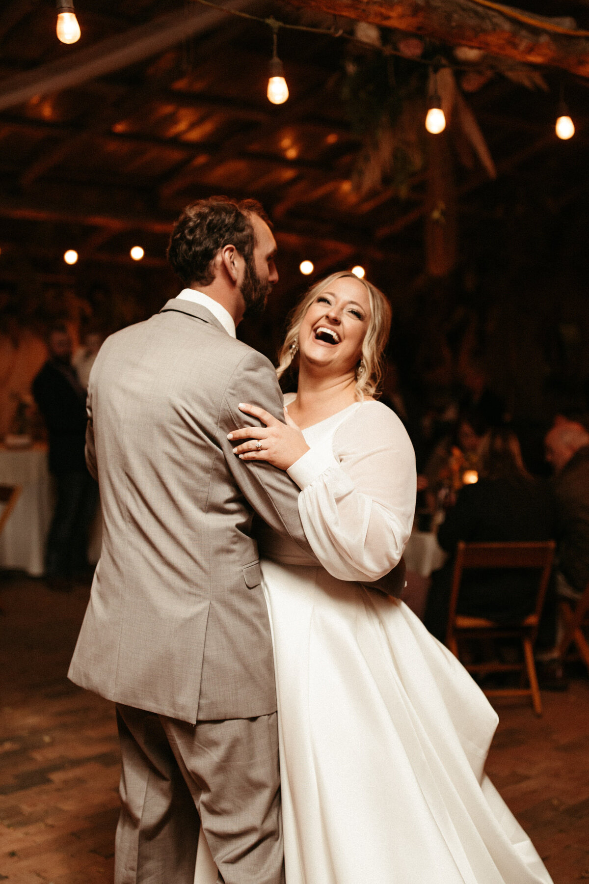Happy bride bursting out in laughter during the first dance with her groom underneath stringed lights