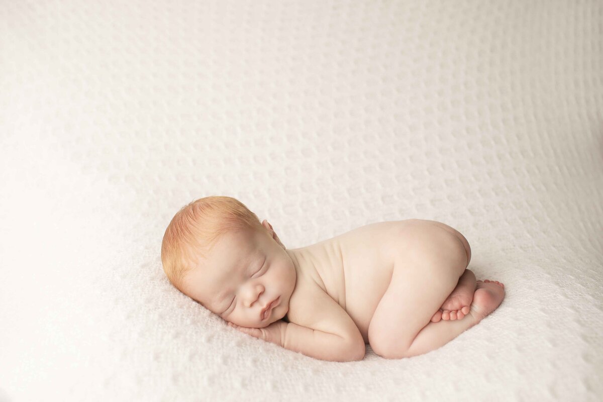 Red-haired newborn baby boy snuggled in a curled-up pose on a smooth backdrop, looking peaceful and content