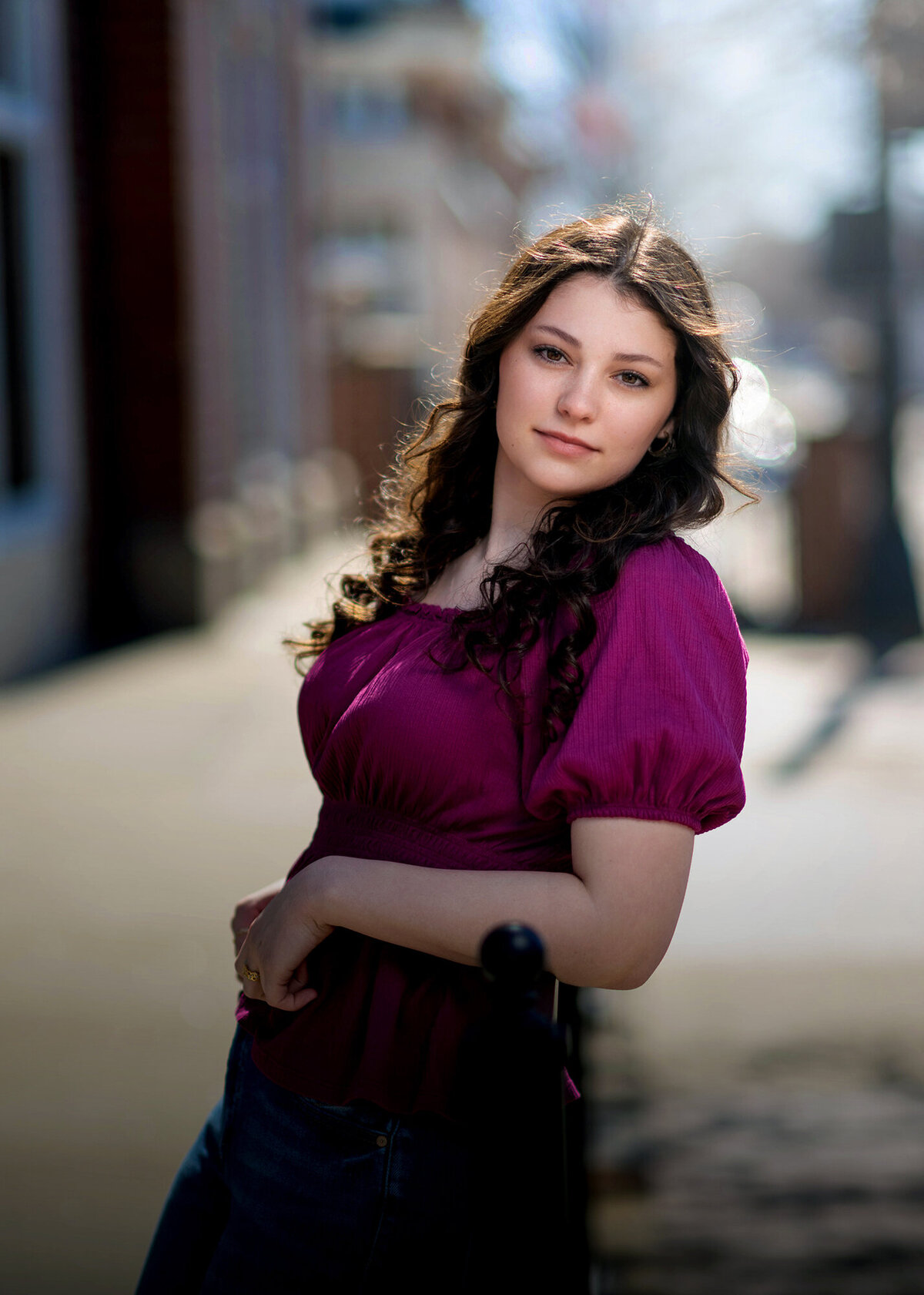 A woman in a purple blouse leans on a railing in the street smiling for her Pella Senior Photographer