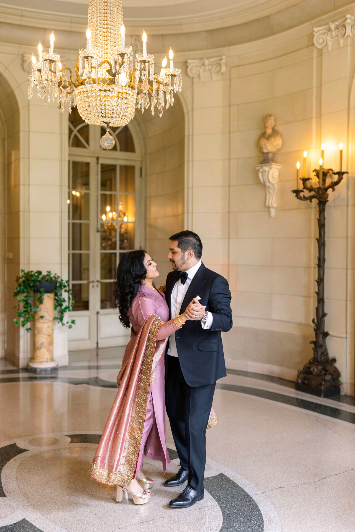 A couple dressed elegantly in formal attire dance together in an ornate room with a large chandelier. The woman wears a pink and gold traditional dress, and the man wears a black tuxedo. They smile at each other, creating a joyful atmosphere.