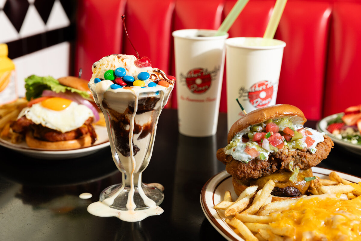 A hero shot of a messy M&M ice cream sundae with a cheese burger and a fried poblano burger and fries.
