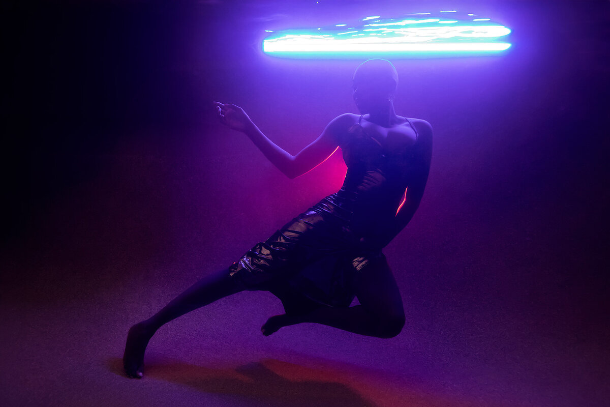 A striking underwater portrait captured by Lindsay Walden Photography featuring a woman in a metallic gown, elegantly posed against a glowing blue and pink neon backdrop. The futuristic lighting creates a vibrant play of shadows and reflections, highlighting the luxurious textures and curves of the gown. An extraordinary display of fine art underwater maternity photography.