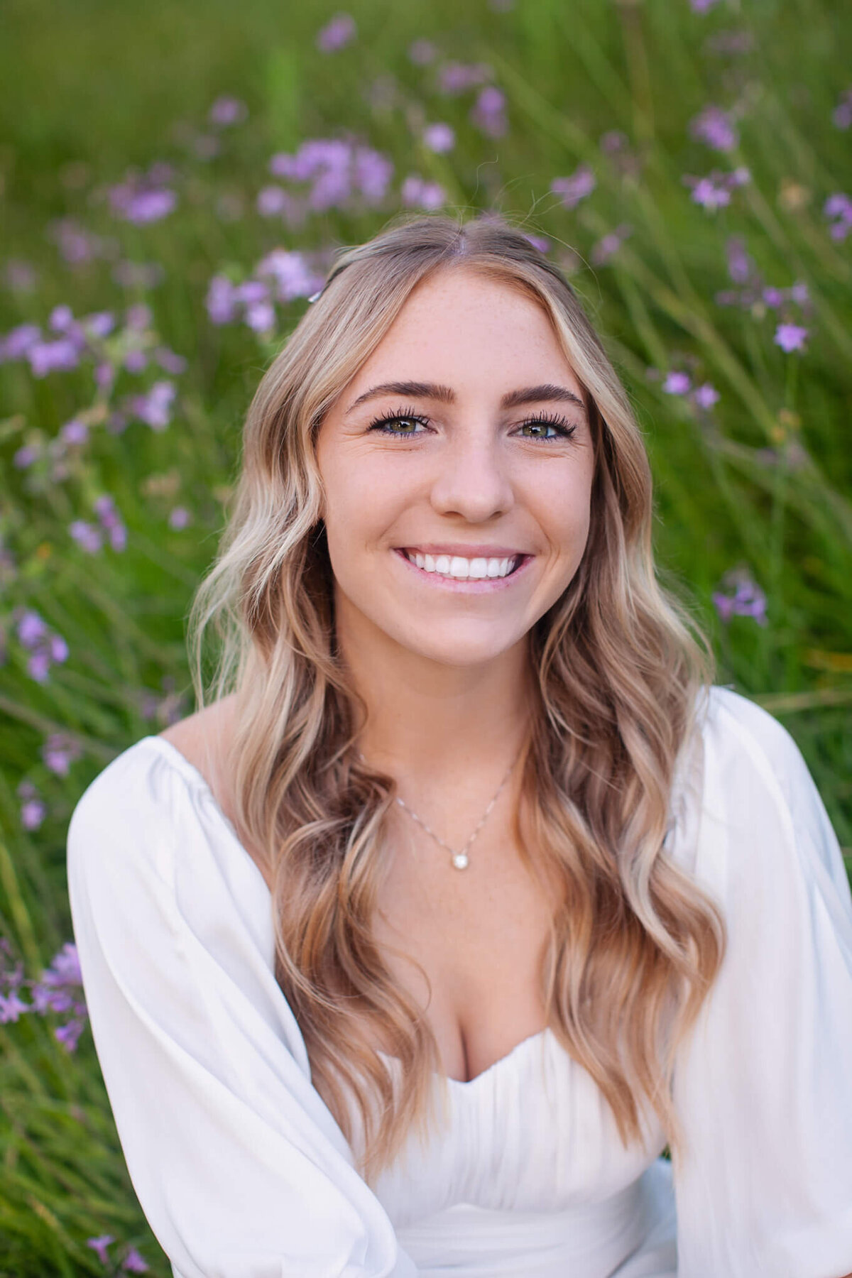 A woman with long wavy hair smiles while seated in a field of green grass and purple flowers. She is wearing a white blouse, and the background is softly focused, emphasizing the peaceful outdoor setting.