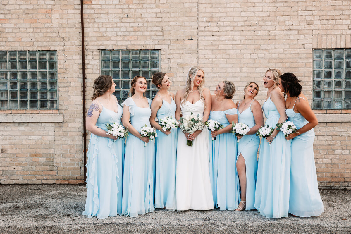 A bride stands holding her bouquet with her bridesmaids and their bouquets by a brick wall