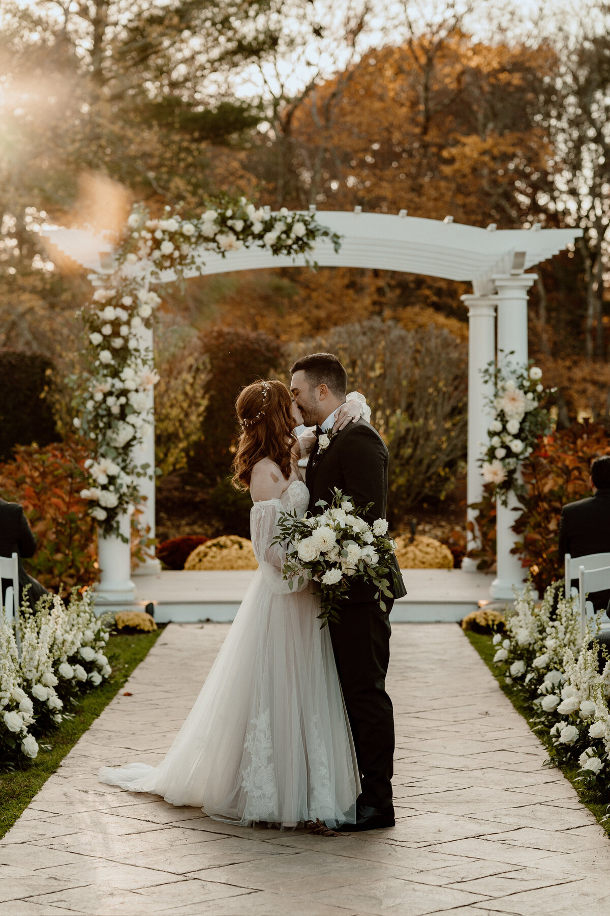 Bride and groom share a romantic kiss under a white floral archway during their outdoor wedding ceremony, surrounded by autumn foliage and decorated with white flowers