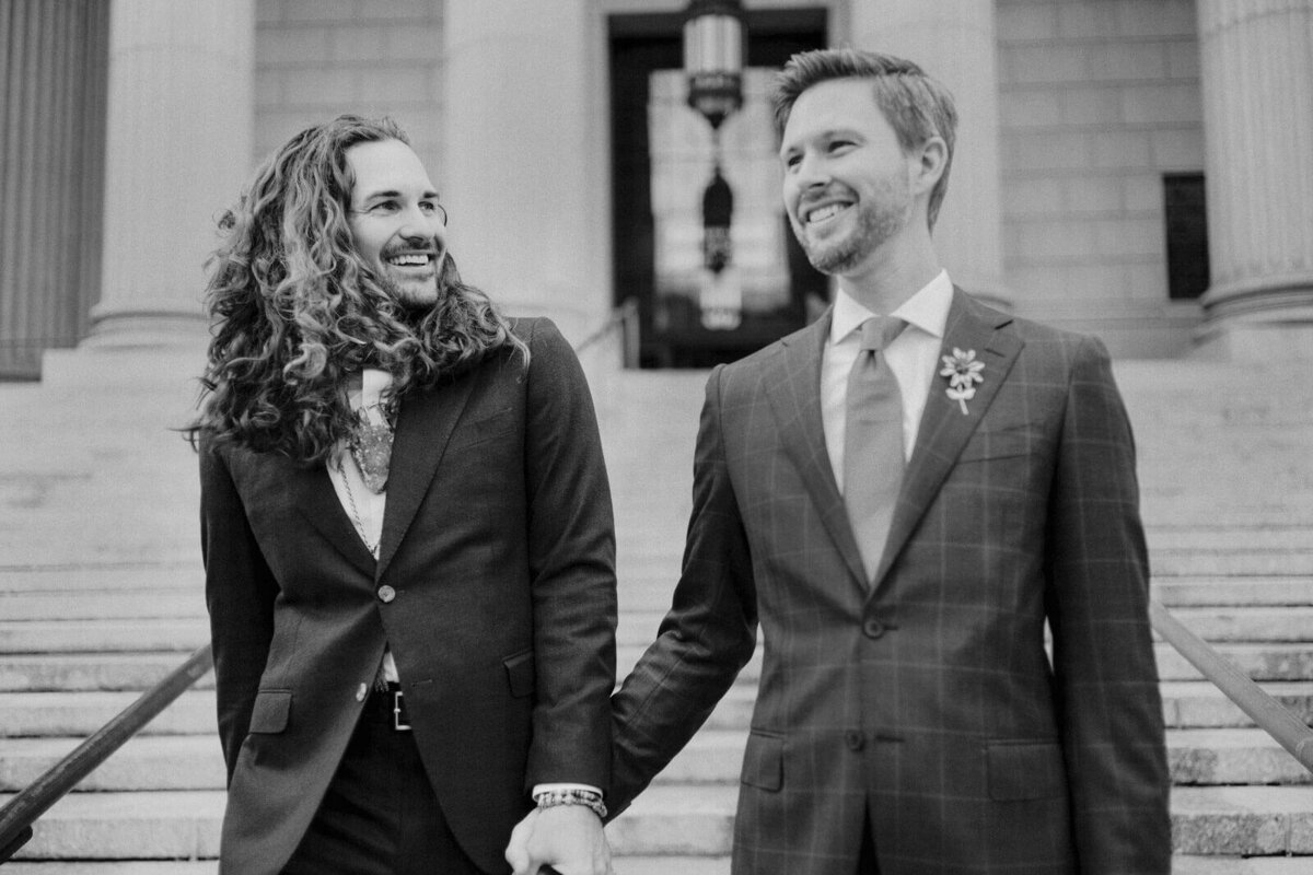 Black and white photo of the two grooms going down the staircase at NYC City Hall. Elopement Image by Jenny Fu Studio