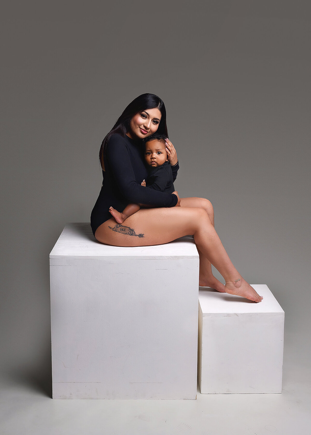 Mom in a black one piece bodysuit with long black hair holding her son who is wearing a black onsie, sitting on white posing blocks in my Phoenix family studio