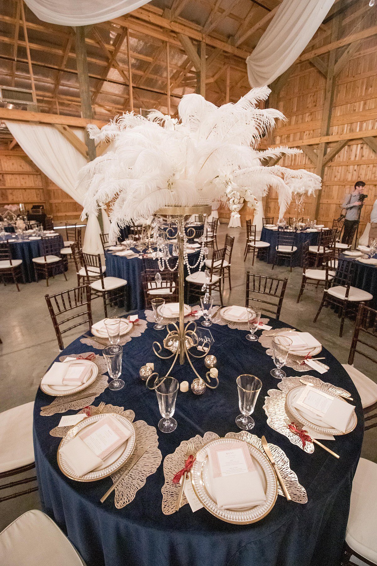 Navy blue velvet table cloths and brown chiavari chairs with white cushions decorate the reception tables at this art deco themed 1920s wedding reception at Saddle Woods Farm. The tables are set with gold ginko leaf chairgers, ivory and gold china plates and menu cards wrapped in champagne linen napkins. The matte gold flatware has a small thank you note tied to each fork and there is a footed crystal water goblet with a gold rim at each place setting. The centerpiece is a tall godl stand with hanging crystals and it is topped with a large plume of white ostrich feathers.