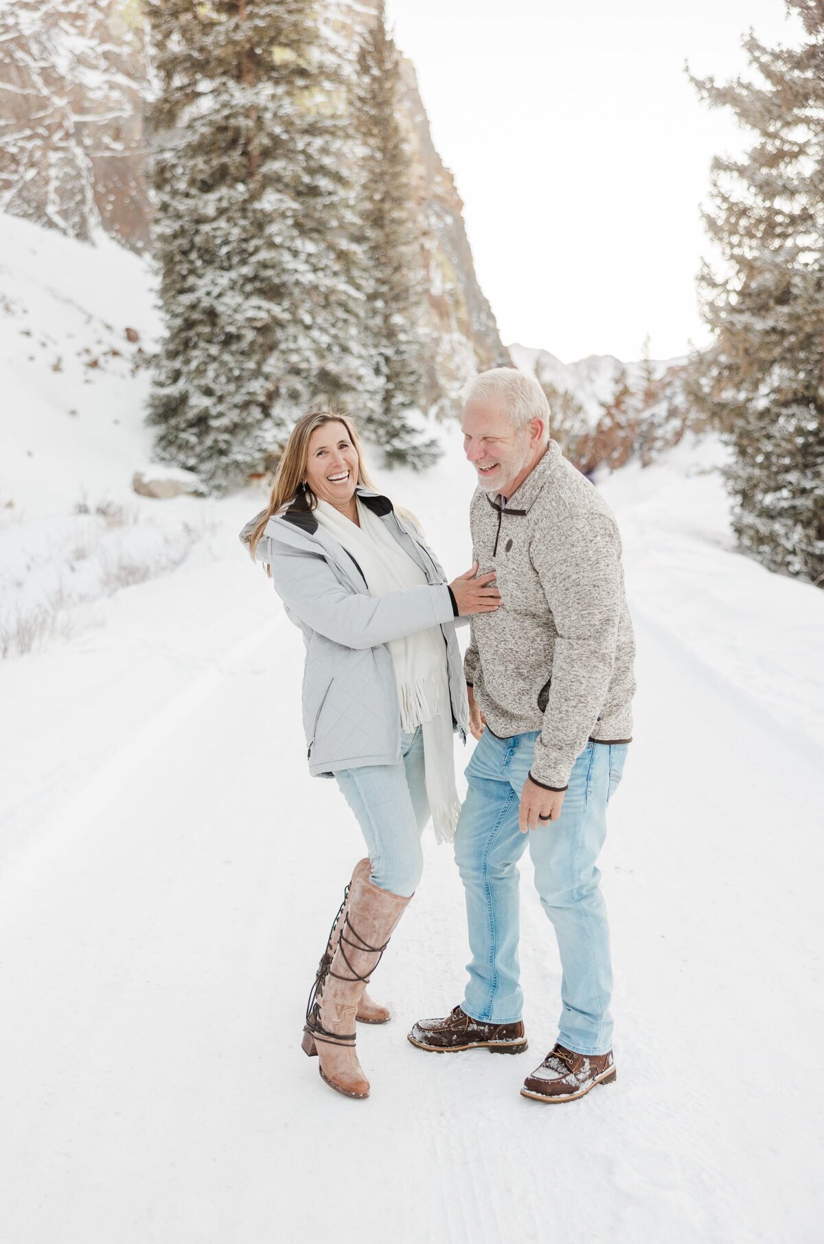 A mom and dad in jeans and sweaters laugh together in a snowy trail
