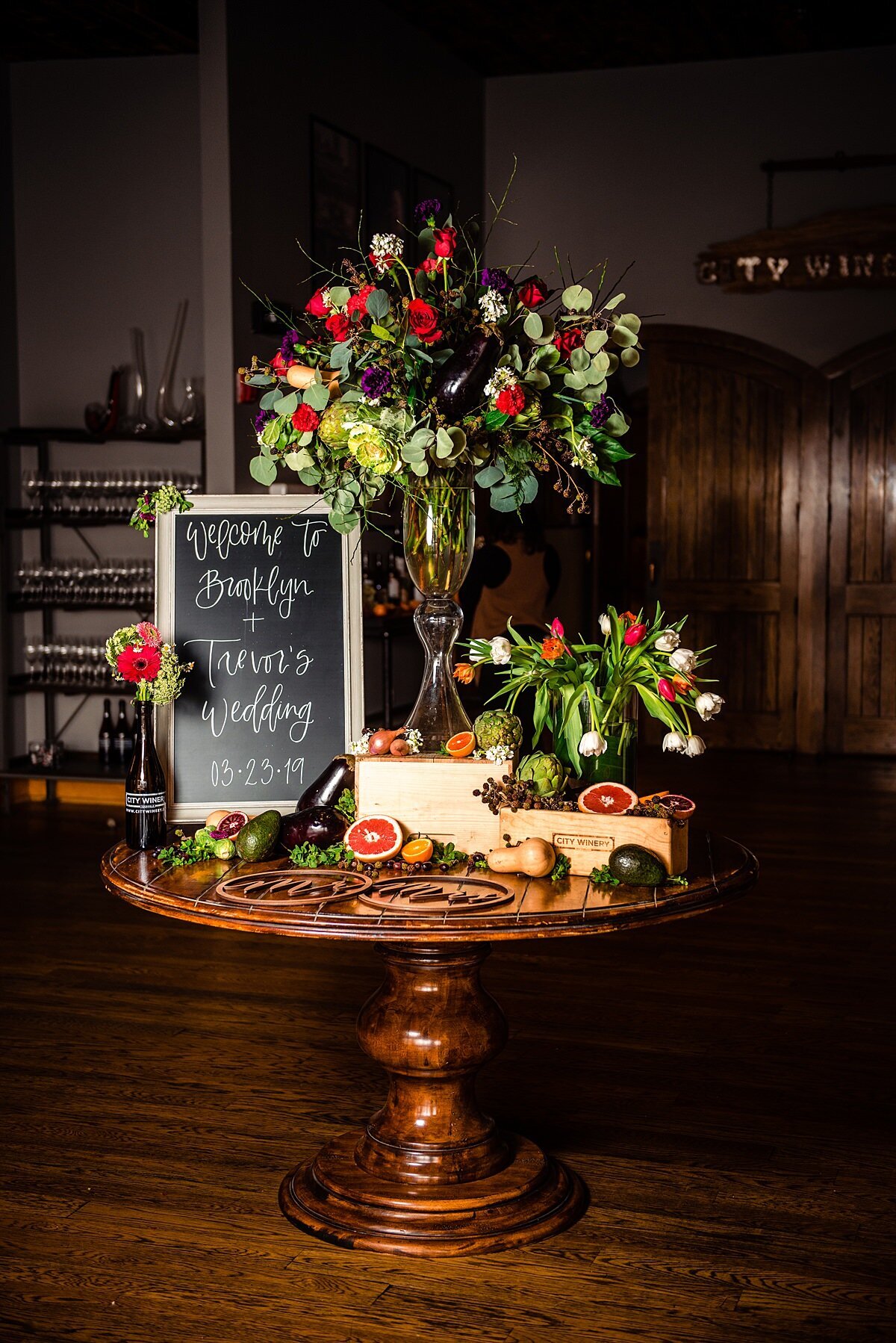 A large dark wood round table is set with a chalkboard sign with white calligraphy sitting next to a tall glass vase brimming with red roses and silver dollar eucalyptus. A low vase sits on the table next to it with white and red tulips. In front of the flowers are two wooden wine boxes with sliced grapefruits, sliced blood oranges, avocados, butternut squash a bottle of City Winery wine and two wooden laser cut signs at City Winery Nashville