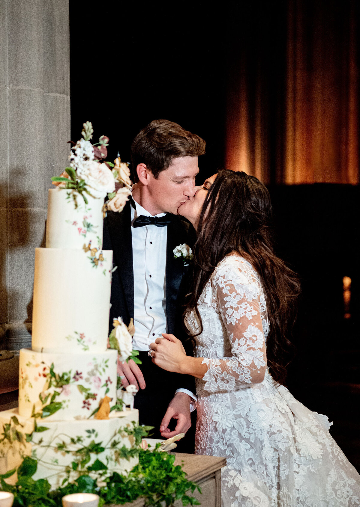 A bride and groom, dressed in a white lace wedding gown and a black suit with a bow tie respectively, share a kiss next to a three-tier wedding cake adorned with flowers and greenery. The background is dimly lit, highlighting the couple's romantic moment.