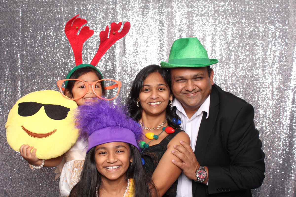 Parents smile as they pose together in a photo booth with their two daughters using props at a wedding