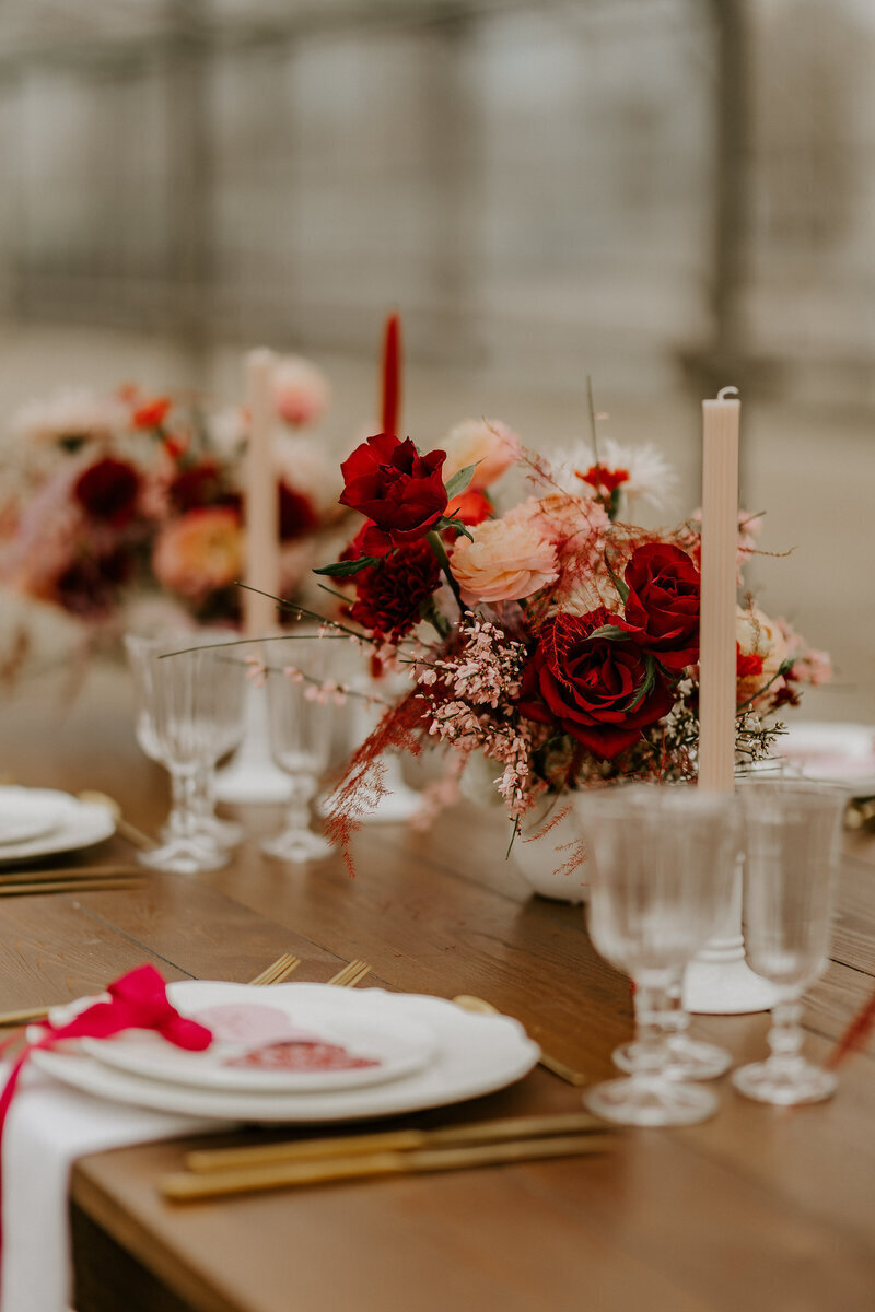 Table en bois décorée de vaisselle, verres, couverts dorés, bougeoirs et bouquets rouges et rose. Arrière plan flou. Séance photo pour le workshop photographie de mariage.