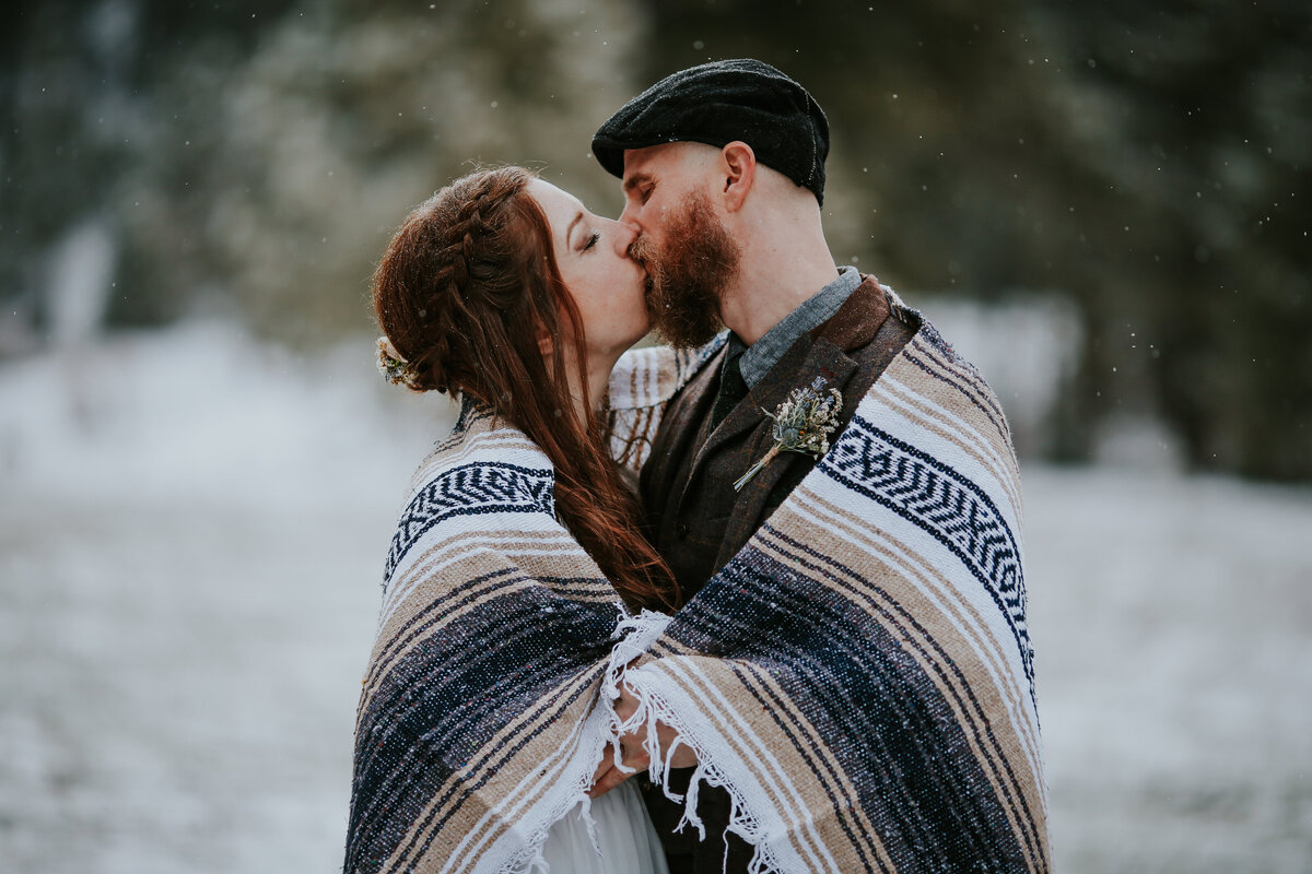 Bride and groom are wrapped in a mexican blanket and kiss as snow lightly falls.