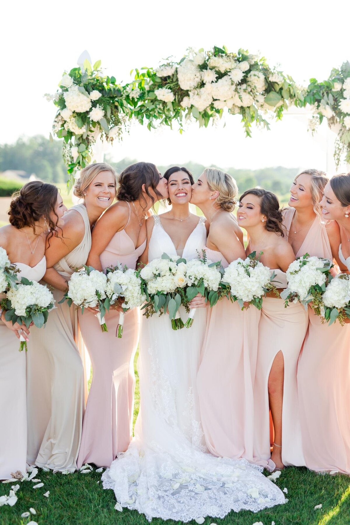 A bride smiling with her bridesmaids under a floral arch, all holding bouquets.