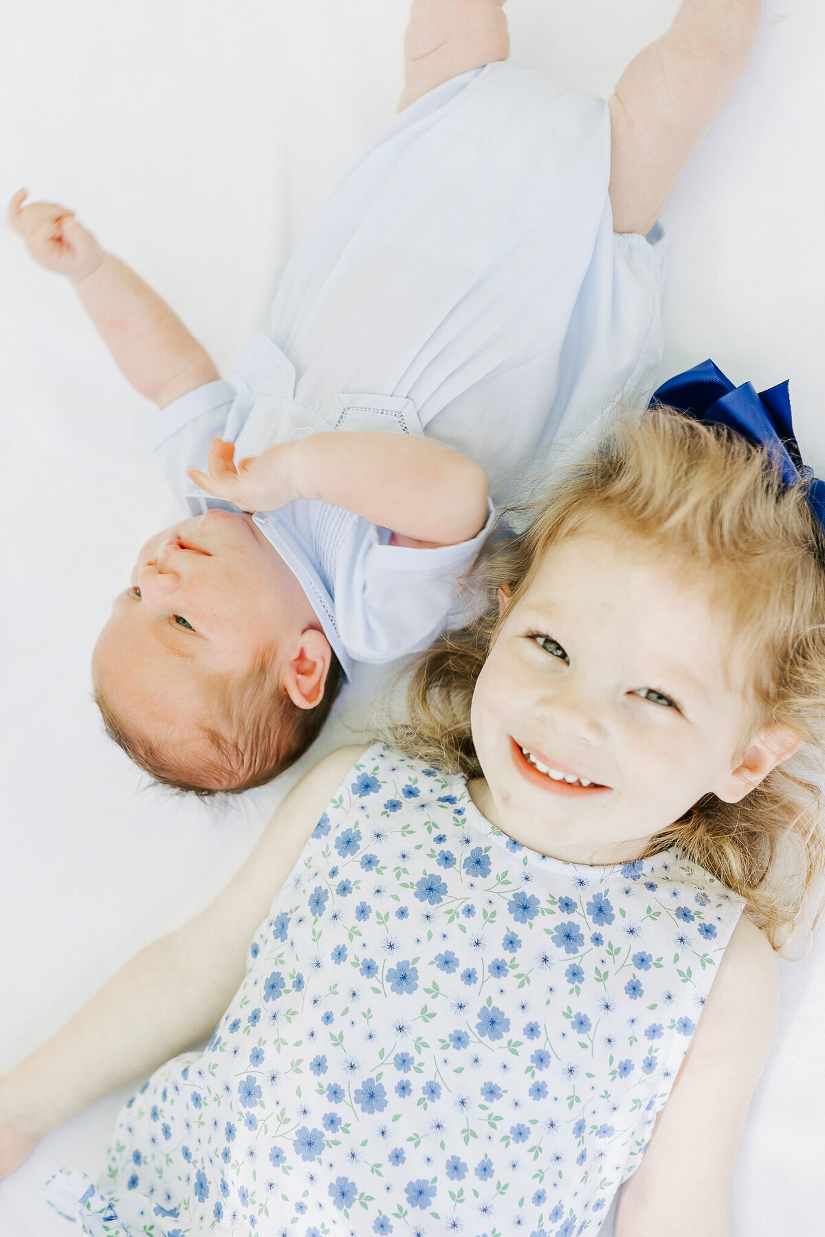 A smiling young girl with a blue bow lies on a white blanket beside her newborn sibling dressed in white.