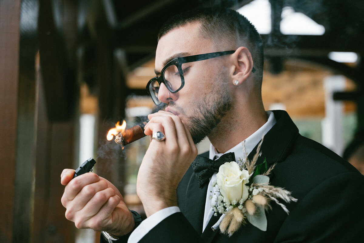 Groom lights cigar prior to his wedding ceremony in his black tux and black-rimed glasses. 