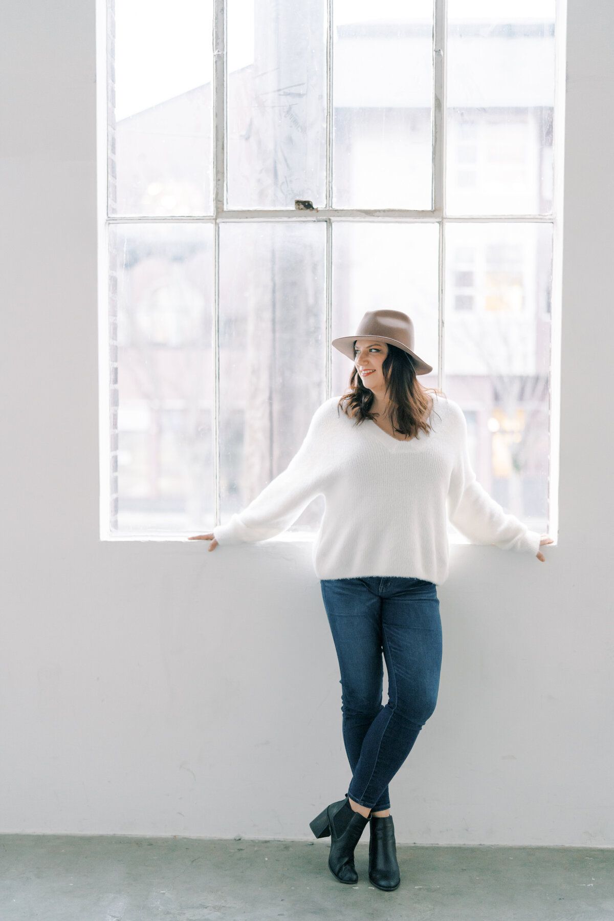 woman in white shirt and tan hat standing in front of large window with her arms stretch out