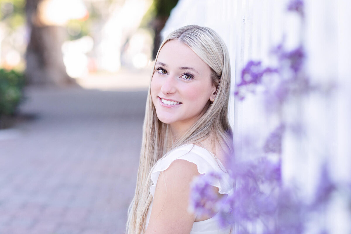 A young woman with long blonde hair, wearing a white dress, leans against a white fence. Purple flowers are in the foreground, and the sidewalk stretches into the blurred background. She smiles gently at the camera.