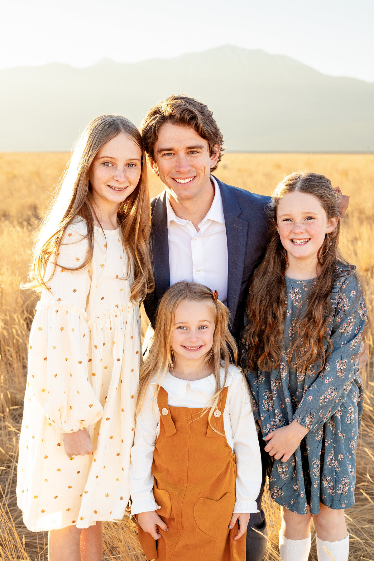 A father kneels in a field, smiling with his three daughters in soft, autumn-colored dresses against a mountainous backdrop.