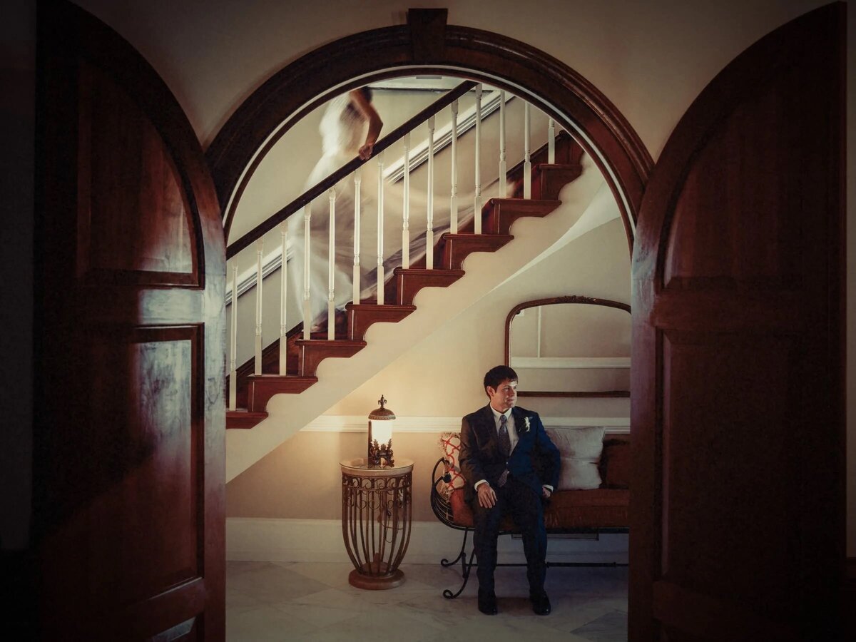 A groom sits contemplatively in a vintage chair, set against a classic staircase with a soft light highlighting his form.
