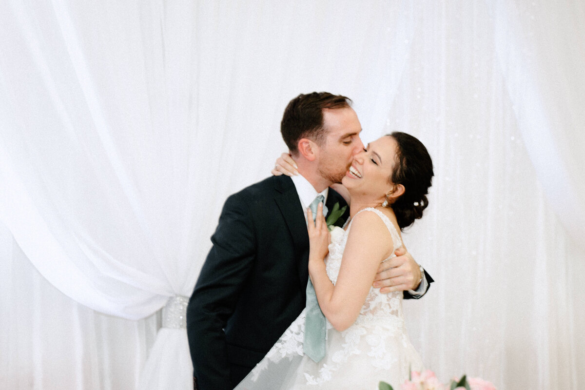 The bride laughing while the groom hugs and kisses her cheek