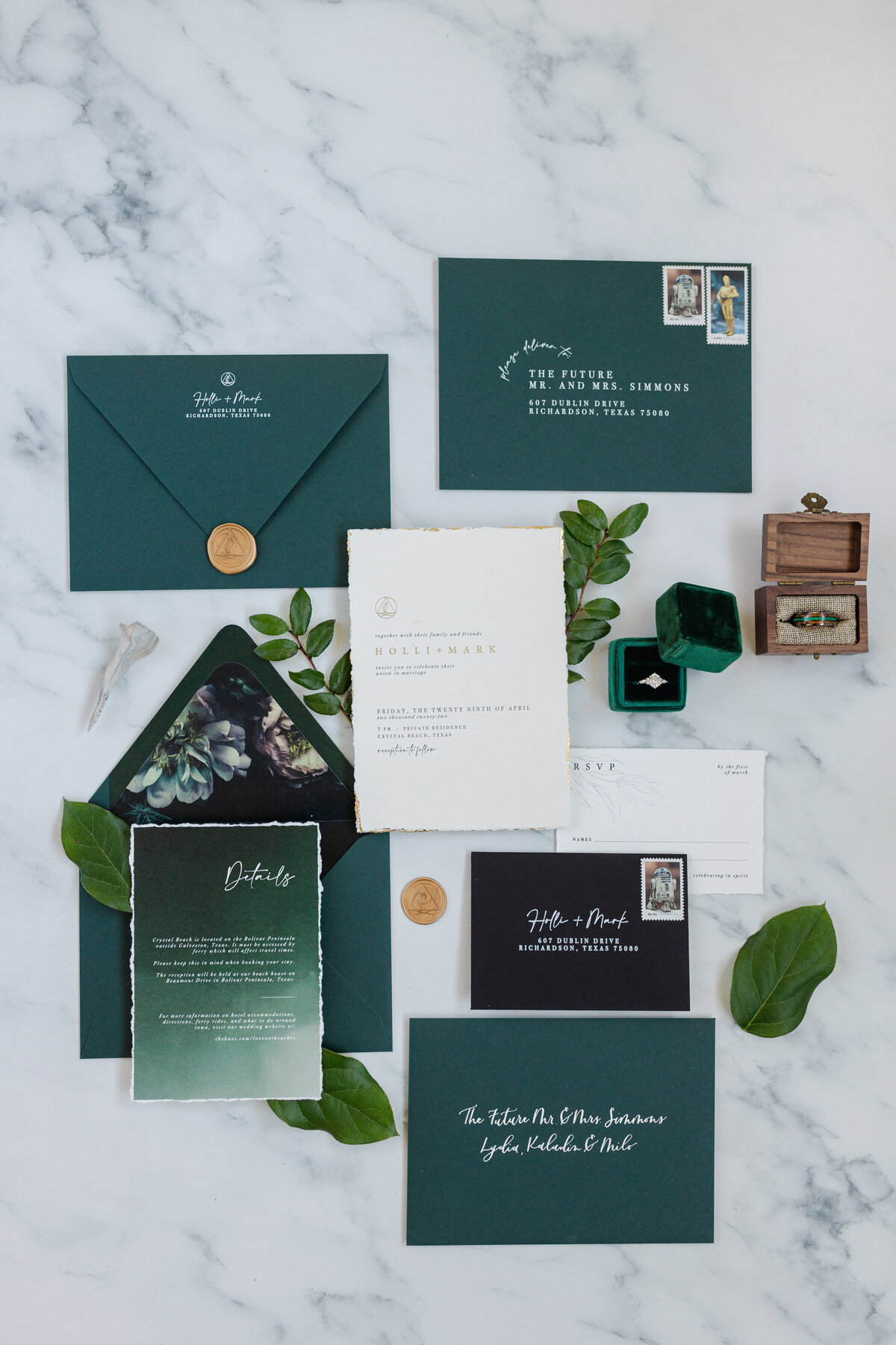 Flat lay detail shot of a bride and groom's invitations, save the dates, envelopes. and wedding rings in their respective boxes at a rental home in Crystal Beach, Texas. The details are resting on a marble floor and our decorated with leaves from the bride's bouquet.