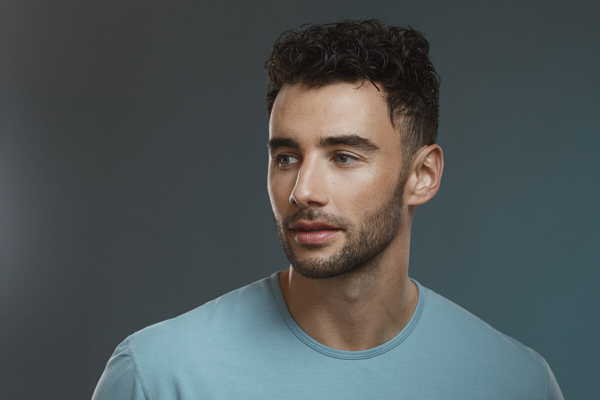 Studio portrait of Brendan Morais in a light blue shirt with a calm expression, photographed against a soft blue background by Ylva Erevall.