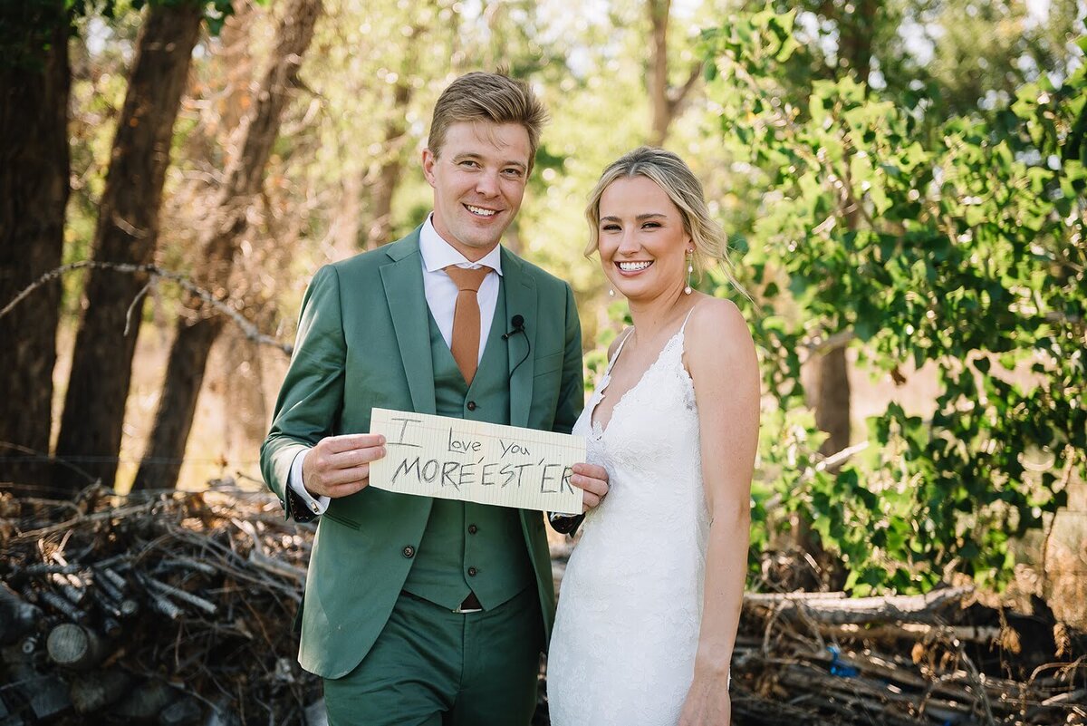 Bride and groom pose with a sign that says I love you more on their wedding day in Colorado .
