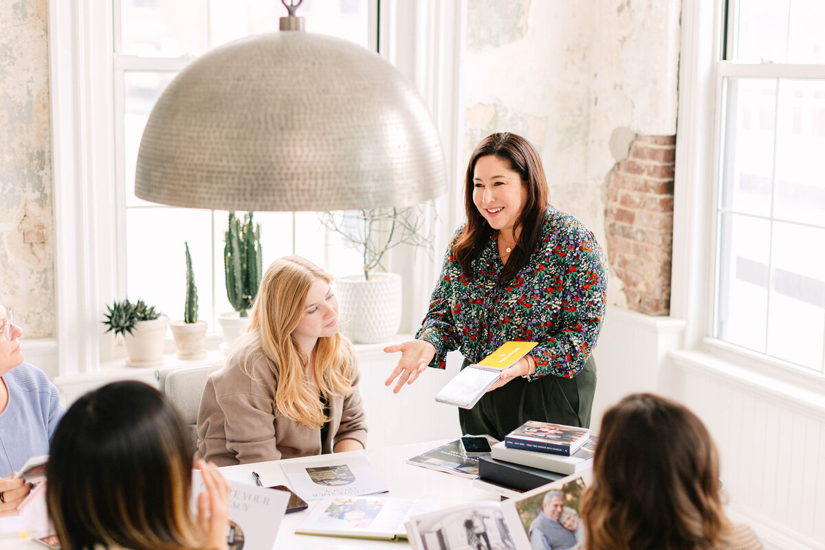 A woman in a floral blouse stands at the head of a table, holding a yellow notebook and gesturing as she speaks to a group of women engaged in discussion. The setting is a bright, modern space with exposed brick and large windows.