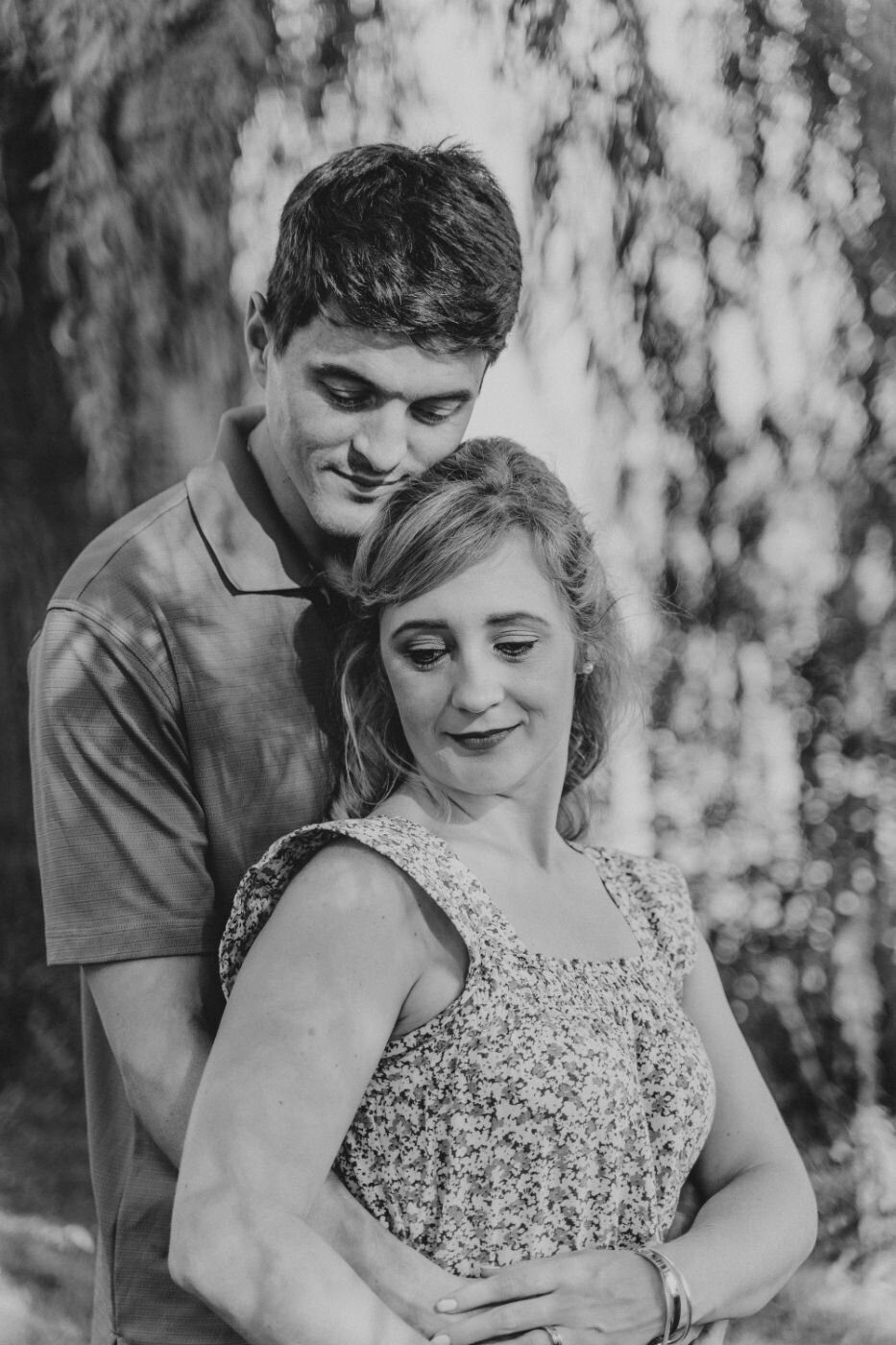 Intimate monochrome portrait of a man gently holding a woman from behind under softly lit trees.