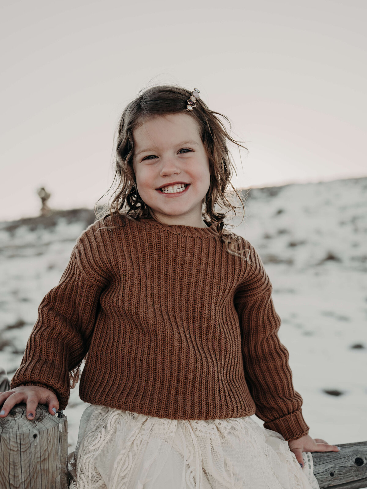 A spunky, three year-old wearing a brown fluffy sweater with wild curly hair grins at the camera while sitting on the top of an old fence post on a family farm in Colorado.