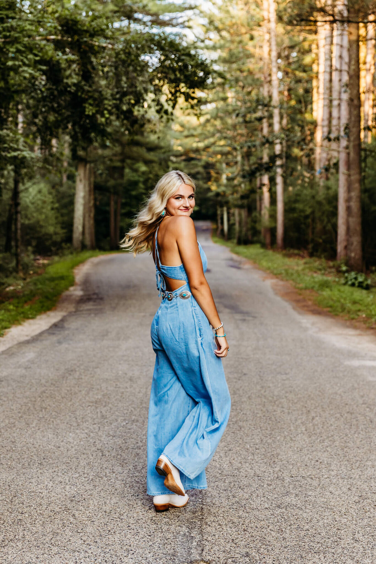 blonde high school girl spinning and looking back as she walks down a backroad that has trees lined on both sides.