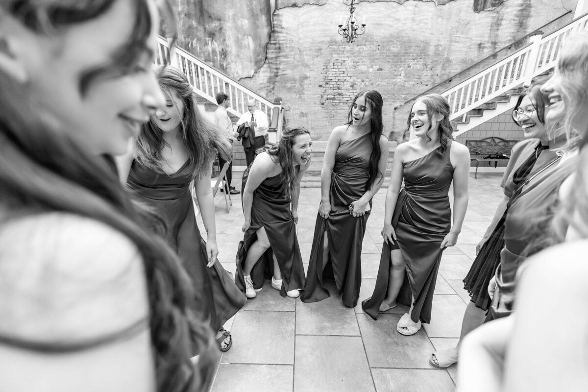 A group of women in formal dresses stand in a circle, laughing and smiling together in a courtyard setting with stairs and brick walls. The image is black and white, capturing a joyful, candid moment.