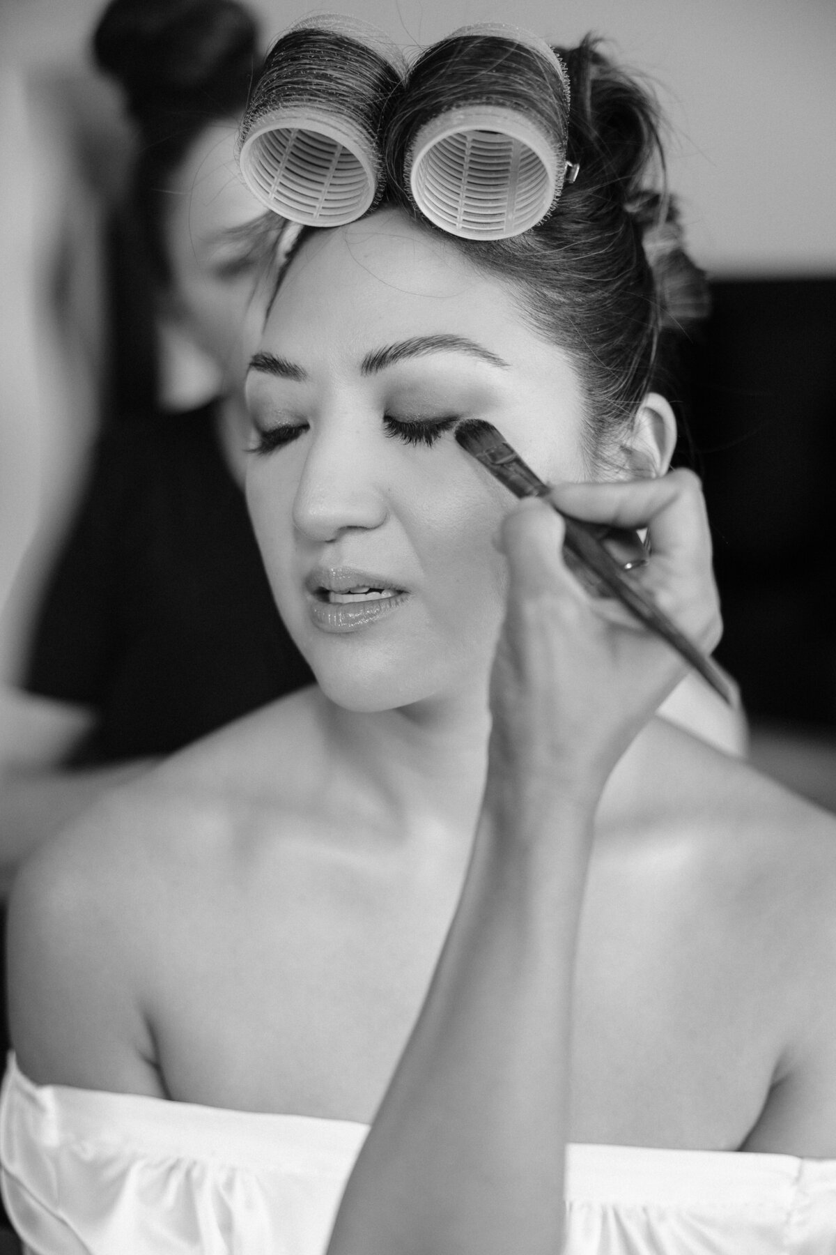 bride with hair in rollers and a hand reaching towards her face with a makeup brush