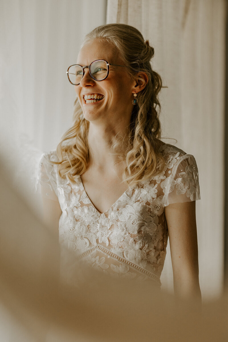 Mariée blonde portant des lunettes, souriante, habillée d'une robe en dentelle blanche posant pour Laura Termeau, photographe professionnelle en vendée.