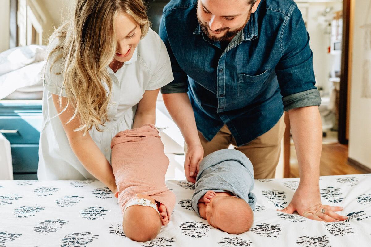A mother and father lean over a bed, carefully placing their swaddled newborn twins on the patterned bedspread.