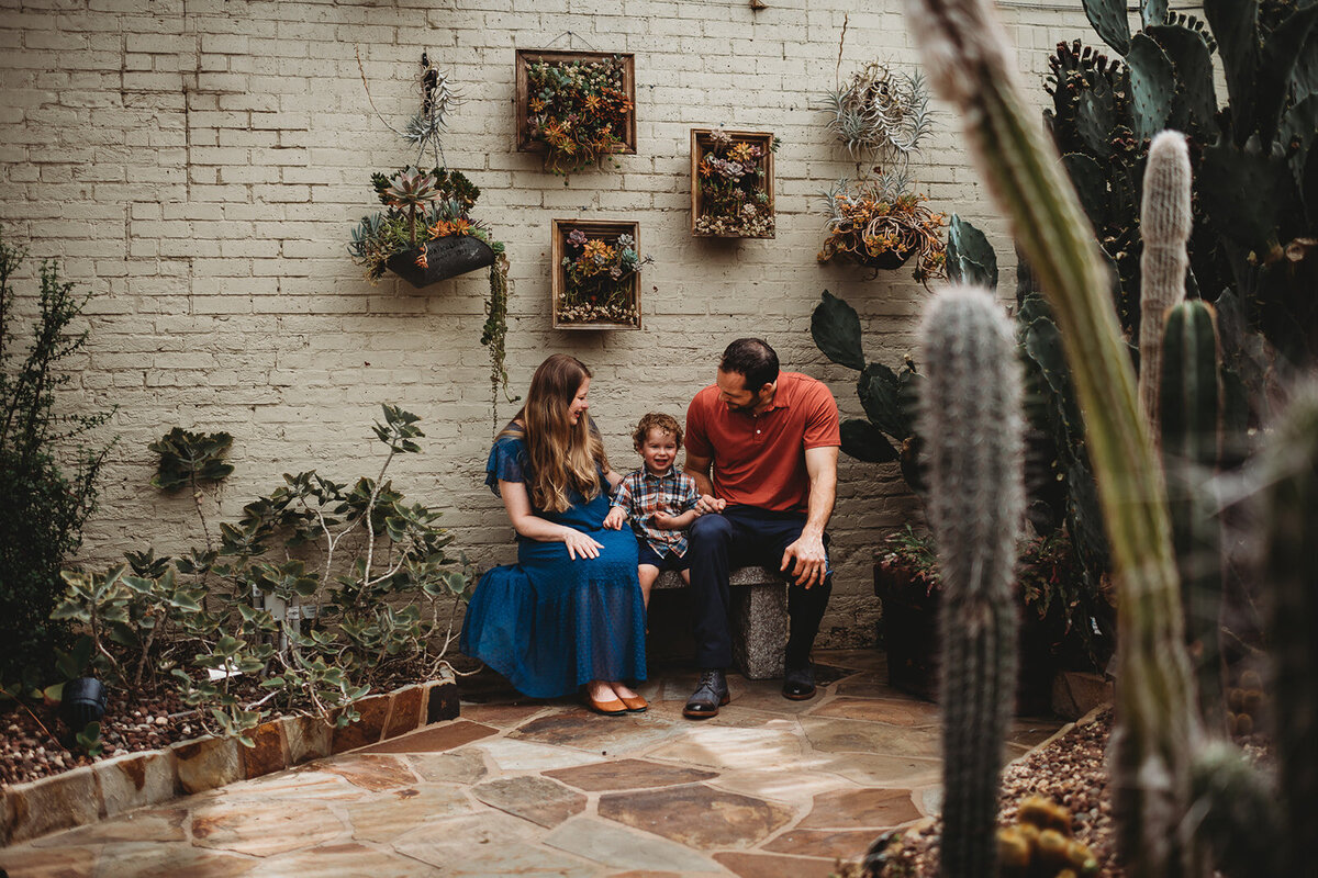 summer family photos with mother and father sitting on a bench in a courtyard with their toddler son captured by Family photographers Maryland