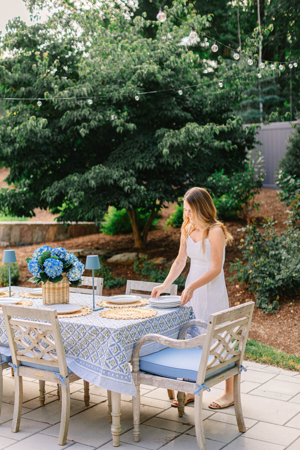 A woman in a white sundress sets plates on an elegantly arranged outdoor dining table with blue accents. The table features a patterned tablecloth, woven placemats, blue glassware, and a wicker basket filled with hydrangeas. String lights hang overhead, and lush greenery surrounds the patio.