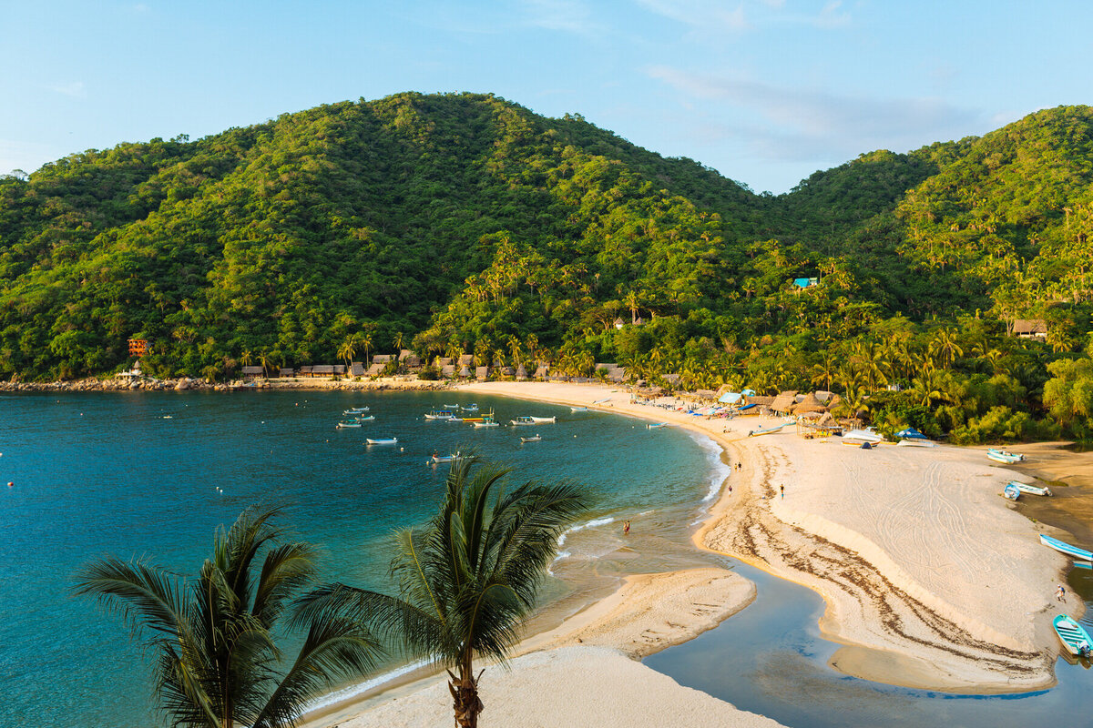 A scenic view of a curved beach bordered by lush green hills, perfect for Caribbean getaways. The sandy shore is lined with palm trees, and small boats float on the calm, blue water. A few people stroll along the beach, and there are thatched-roof structures near the trees.