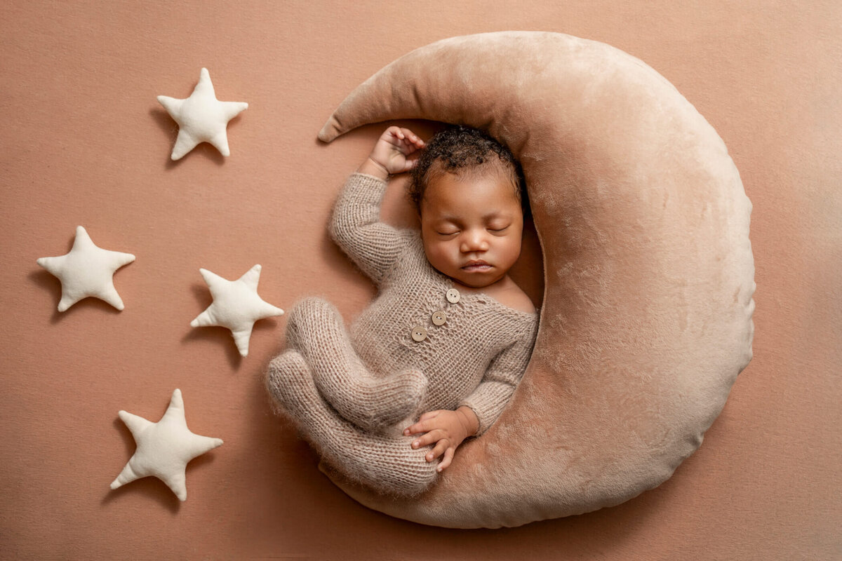 A sleeping newborn baby wearing a knitted beige onesie, lying on a plush crescent moon pillow. The baby is surrounded by four soft star pillows on a matching beige background.