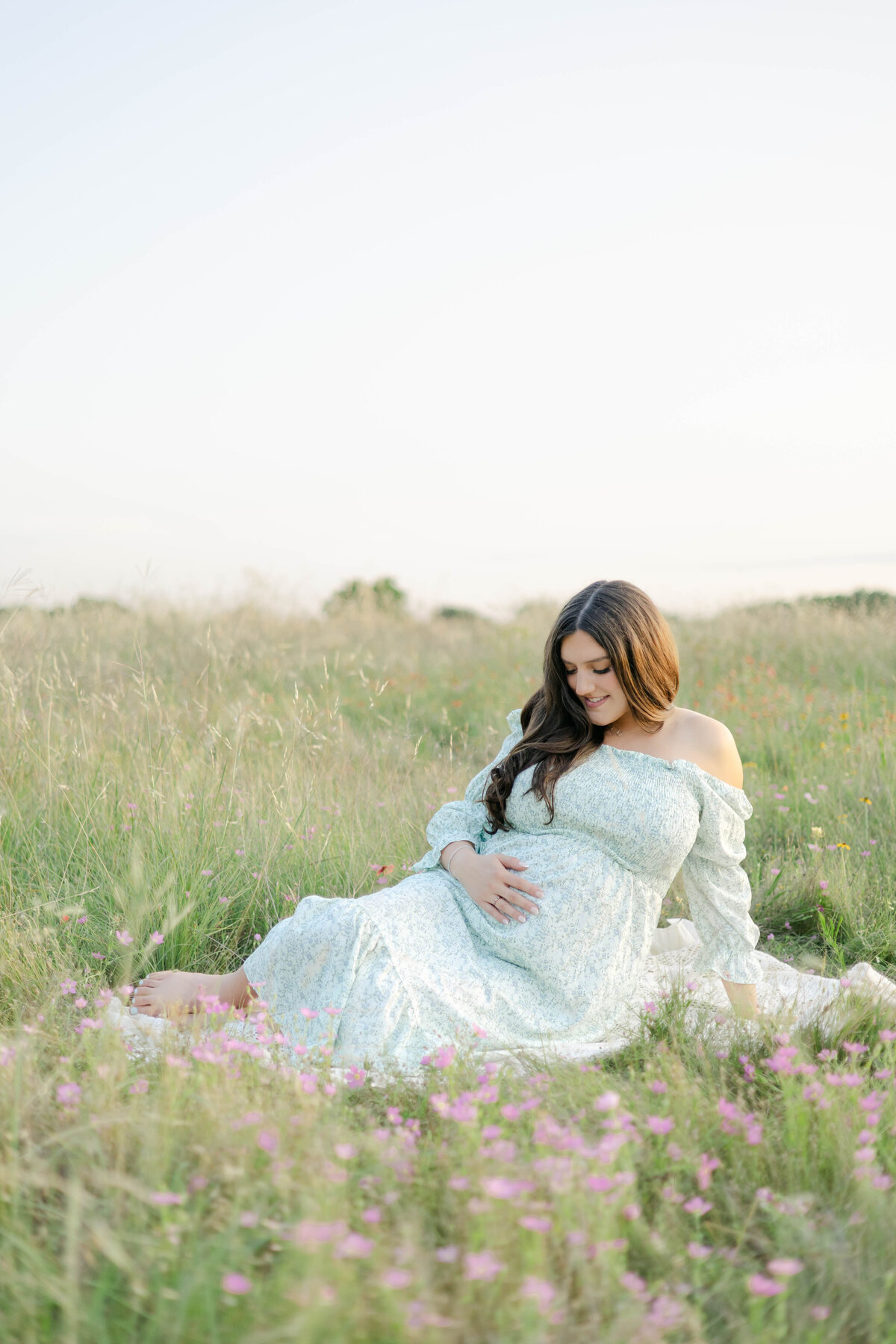 pregnant woman holding her belly sitting in a flower field in spring in dallas texas wearing a green off shoulder dress