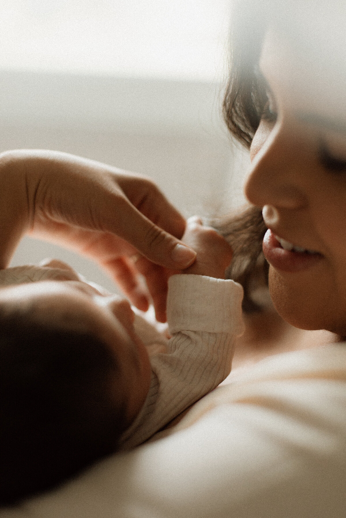 Mother smiling softly as her newborn gently grasps a lock of her hair, bathed in soft window light.