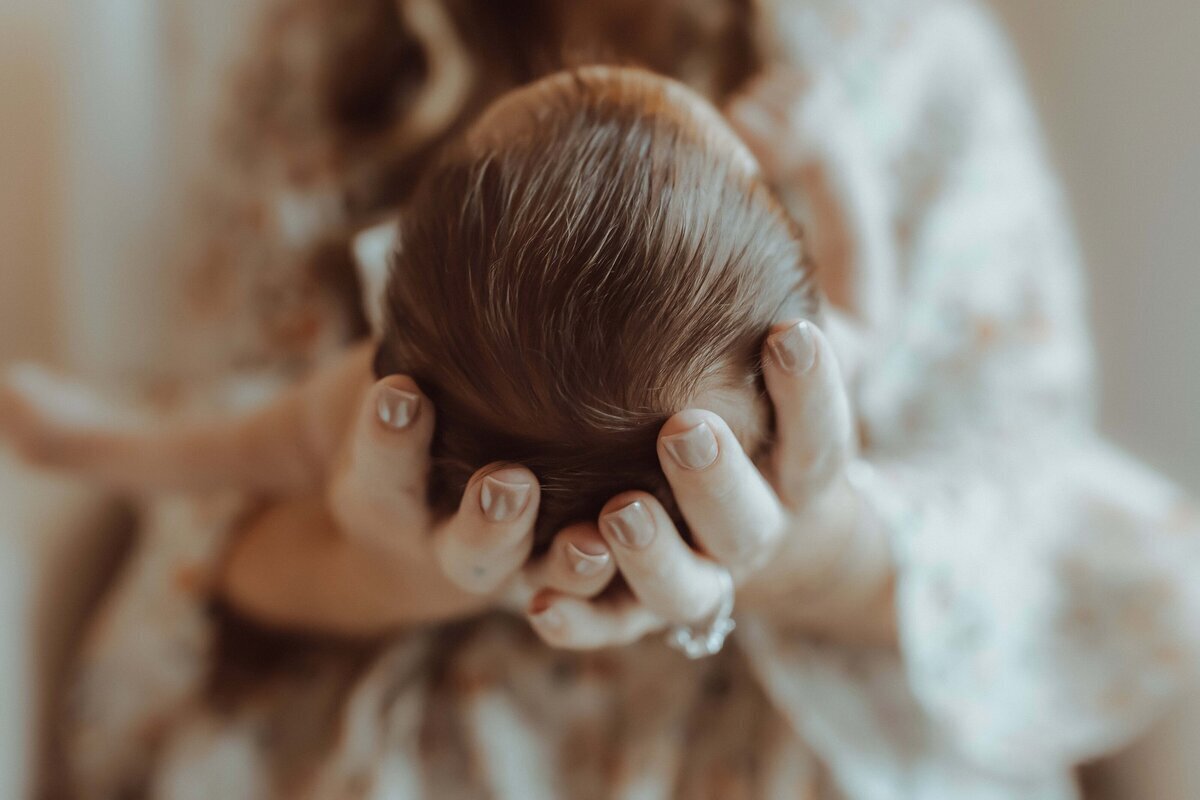 A person cradles a newborn's head gently in their hands, focusing on the baby's fine hair and the tenderness of the moment.