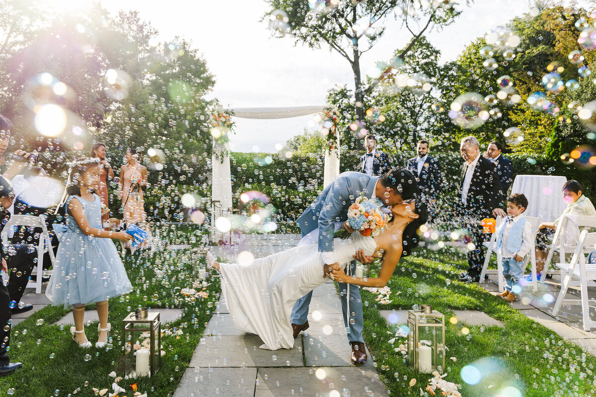 Couple kissing at the end of a garden wedding ceremony while guests blow bubbles around them