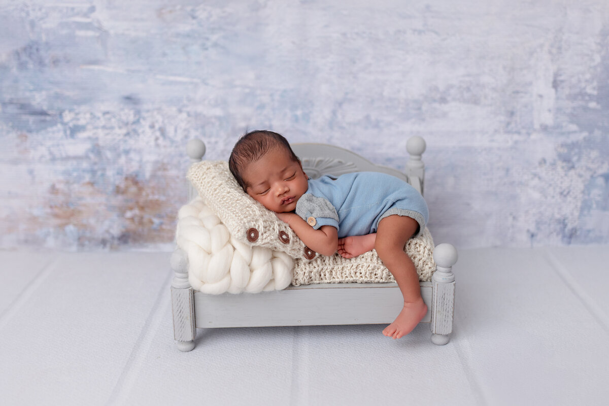 Newborn boy lounging on a small white bed with a soft blue textured backdrop.